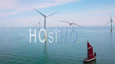 A Flat Bottomed Sailing Barge Sailboat Moves Up The Thames River Estuary In England Amidst Numerous Wind Turbine Windmills - Video Drone Footage