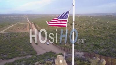 The American Flag Flies Over The U.S. Mexico Border Wall In The California Desert As A Border Patrol Vehicle Passes Below - Video Drone Footage